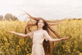 Young beautiful girl with long hair flying in the wind against the background of rapeseed field. Breeze playing with girl`s hair