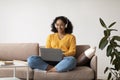 Young happy black woman in headphones using laptop pc, working online, sitting on couch at home, copy space Royalty Free Stock Photo