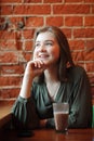 Young happy blond woman in green blouse sitting near window against red brick wall at the cafe with c cocoa glass