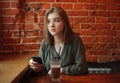 Young happy blond woman in green blouse sitting near window against red brick wall at the cafe with c cocoa glass texting on smart Royalty Free Stock Photo