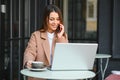 Young happy business lady looks at smartphone screen with toothy smile while sitting outside on terrace with laptop and coffee, Royalty Free Stock Photo