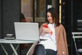 Young happy business lady looks at smartphone screen with toothy smile while sitting outside on terrace with laptop and coffee, Royalty Free Stock Photo