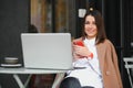 Young happy business lady looks at smartphone screen with toothy smile while sitting outside on terrace with laptop and coffee, Royalty Free Stock Photo