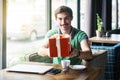 Young happy businessman in green t-shirt sitting and holding and giving big red gift box. looking at camera with toothy smile Royalty Free Stock Photo