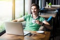 Young happy businessman in green t-shirt sitting and looking ot laptop display with smile, holding and drinking cup of coffee, Royalty Free Stock Photo