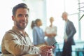 Young happy businessman sitting in the modern office, holding smartphone and smiling at camera while his colleagues Royalty Free Stock Photo