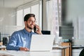 Young happy busy latin businessman talking on cell phone using laptop in office. Royalty Free Stock Photo