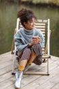 Young happy carefree african american woman with cup of hot tea relaxing near pond in autumn time, mixed-race female in warm Royalty Free Stock Photo