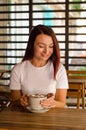 Young caucasian woman sits at wooden table with cup, on terrace of cafe with striped background and flowers Royalty Free Stock Photo