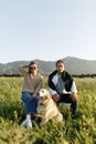 Young happy couple with dog, sitting on chairs in feather grass field on summer sunset day, looking forward in camera Royalty Free Stock Photo