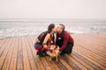 Young happy couple kissing each other and playing with dog on the rainy berth in autumn. Sea background Royalty Free Stock Photo