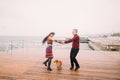 Young happy couple walking with dog and having fun on the rainy berth in autumn. Sea background Royalty Free Stock Photo