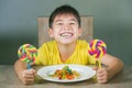 Young happy and excited male kid smiling cheerful eating dish full of candy and lollipop sitting at table isolated on grey Royalty Free Stock Photo