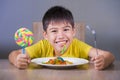 Young happy and excited male kid smiling cheerful eating dish full of candy and lollipop sitting at table isolated on grey Royalty Free Stock Photo