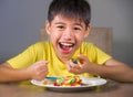 Young happy and excited male kid smiling cheerful eating dish full of candy and lollipop sitting at table isolated on grey Royalty Free Stock Photo