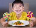 Young happy and excited male kid smiling cheerful eating dish full of candy and lollipop sitting at table isolated on grey Royalty Free Stock Photo