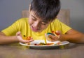 Young happy and excited male kid smiling cheerful eating dish full of candy and lollipop sitting at table isolated on grey Royalty Free Stock Photo