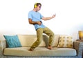 Young happy and excited man jumping on sofa couch listening to music with mobile phone and headphones playing air guitar crazy hav Royalty Free Stock Photo