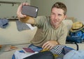 Young happy and excited man taking selfie portrait with mobile phone camera while packing travel suitcase and preparing mess cloth Royalty Free Stock Photo