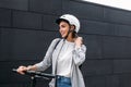 Young happy female adjusting strap of a backpack while standing at wall on electric scooter. Businesswoman with cycling helmet