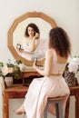 Young happy girl with curly hair does makeup in front of a vintage mirror. A beautiful woman in a pink dress paints her eyes Royalty Free Stock Photo