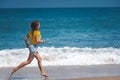 Young happy girl in a yellow T-shirt and shorts runs barefoot along the ocean, sea horizon and white waves on a sunny day, Royalty Free Stock Photo