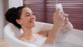 Young happy hispanic woman lying in hot comfortable foam luxurious bath relaxed girl typing sms on phone in bathroom Royalty Free Stock Photo