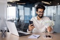 Young happy Indian man sitting in the office at the desk, holding the phone and looking at a fan of cash money Royalty Free Stock Photo