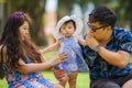 Young happy loving Asian Korean parents couple enjoying together sweet daughter baby girl sitting on grass at green city park in Royalty Free Stock Photo