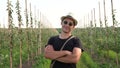 Young happy man in black t-shirt, sunglasses and hat stands with corssed hands among his apple garden and looking at the