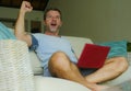 Young happy man sitting at home sofa couch relaxed using internet working on laptop computer gesturing victory as winner excited a