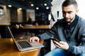 Young happy man smiling and holding a smartphone sitting with laptop in cafe. Portrait man reading a message with smartphone in ca Royalty Free Stock Photo