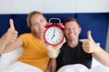 Young and happy man and woman holding red alarm clock and thumbs up while lying in bedroom Royalty Free Stock Photo