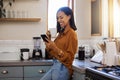 Young happy mixed race woman drinking a cup of coffee while using her phone alone in the morning in the kitchen. One Royalty Free Stock Photo