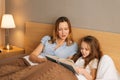 Young happy mother and adorable daughter reading children book together lying in bed before going to sleep Royalty Free Stock Photo