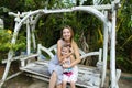 Young happy mother with daughter riding on swing in exotic garden, palms in background.