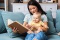 Young happy mother reading a fairytale story book to her baby. Mommy and kid sitting on sofa at home enjoying in imagination.