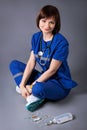 Young happy nurse sitting on the floor posing on gray background. Medical dropper and a stethoscope of a medic Royalty Free Stock Photo