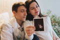 Young happy pregnant woman holding pregnancy ultrasound, showing sonogram picture to pleasantly surprised husband crying from