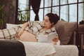 Young happy pregnant woman in pajama reading book and touching belly gently while relaxing at home Royalty Free Stock Photo