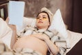 Young happy pregnant woman in pajama reading book and touching belly gently while relaxing at home Royalty Free Stock Photo