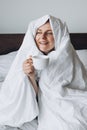 Young happy redhead woman with cup of coffee sitting in bed under blanket at home bedroom. comfort or home weeekend Royalty Free Stock Photo