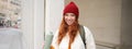 Young happy redhead woman in red hat, drinking from thermos, warming up with hot drink in her flask while walking around Royalty Free Stock Photo