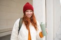 Young happy redhead woman in red hat, drinking from thermos, warming up with hot drink in her flask while walking around Royalty Free Stock Photo