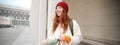 Young happy redhead woman in red hat, drinking from thermos, warming up with hot drink in her flask while walking around Royalty Free Stock Photo