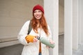 Young happy redhead woman in red hat, drinking from thermos, warming up with hot drink in her flask while walking around Royalty Free Stock Photo