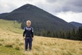 Young happy smiling child boy with backpack standing in mountain grassy valley on background of summer landscape, woody mountain