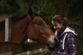Young happy smiling woman horse owner stroking horse head at wooden fence in forest