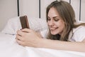 Young happy smiling woman using her phone while lying in bed in the morning. Awakening. Girl reads good news, social Royalty Free Stock Photo