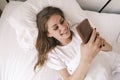 Young happy smiling woman using her phone while lying in bed in the morning. Awakening. Girl reads good news, social Royalty Free Stock Photo